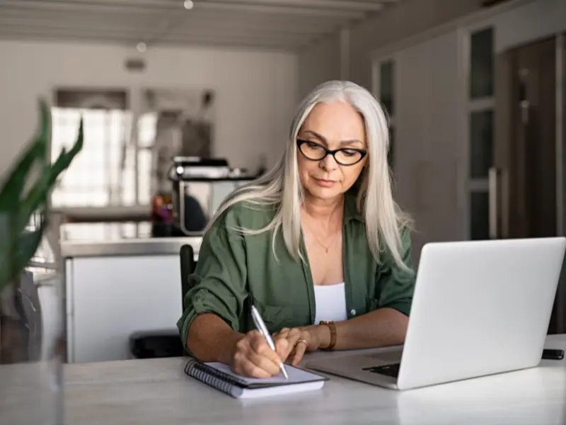 A starting freelance writer with long gray hair sits at a desk, writing in a notebook next to a laptop, in a modern, bright workspace.