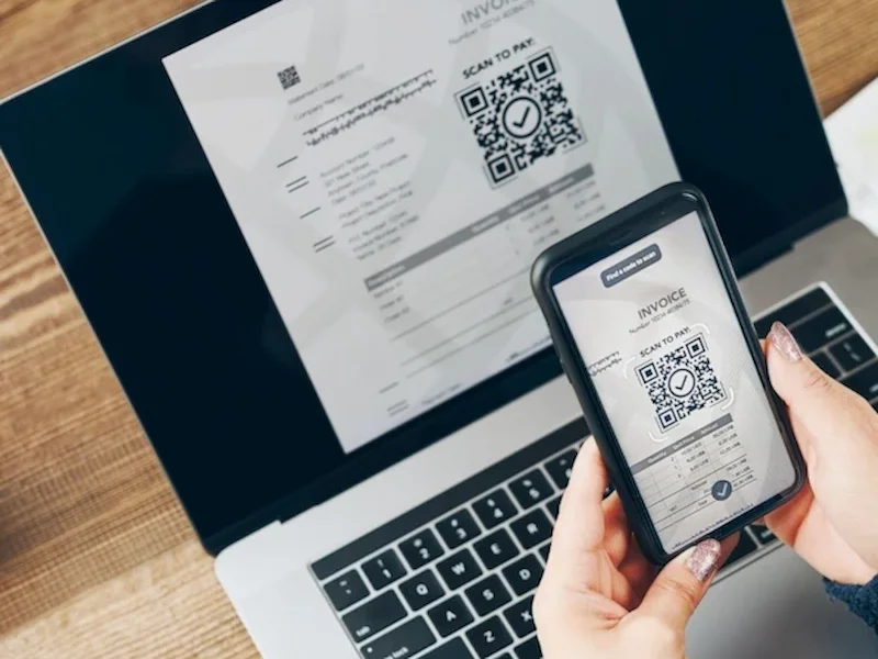 Explore secure payment methods online as a person scans a QR code from an invoice using their smartphone, with a laptop and coffee cup nearby