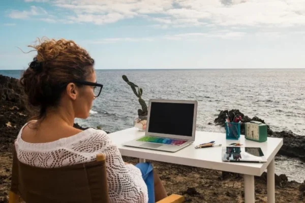 A Person working on a laptop by the ocean with notepad and pens on a rocky terrain surrounded by plants, representing the Freelance2Retire lifestyle of remote work and earning money for retirement.
