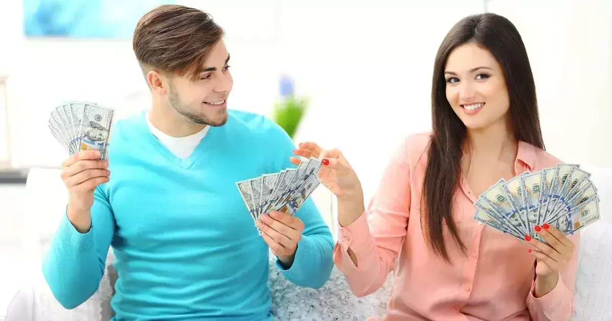 A man in a blue sweater and a woman in a pink blouse joyfully display wads of cash while sitting on a couch.