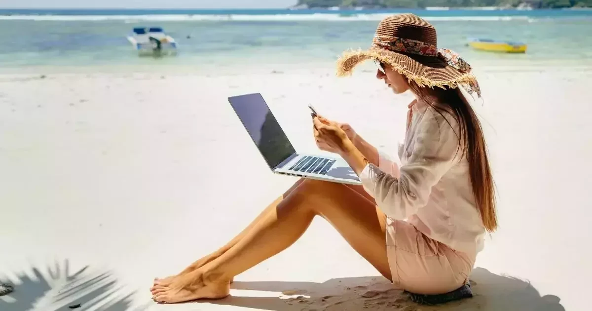 A woman sits on the beach with a laptop and phone, enjoying the sun and ocean view while working remotely.